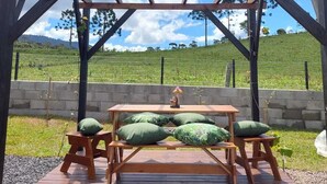 Eine Pergola aus Holz mit Picknicktisch und Bänken, vor einer Wiese und blauem Himmel.