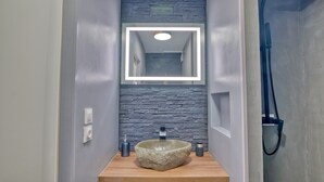 A modern bathroom with a stone-textured wall, a wooden vanity, a stone sink, and a built-in mirror with lighting. - Le Grain de Sable (Agde)