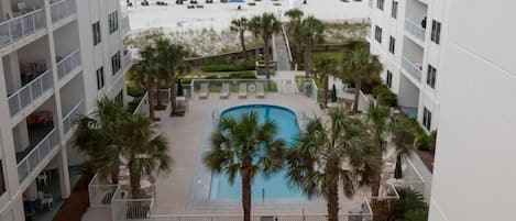 A pool area with a view of the beach, surrounded by palm trees and a white building.