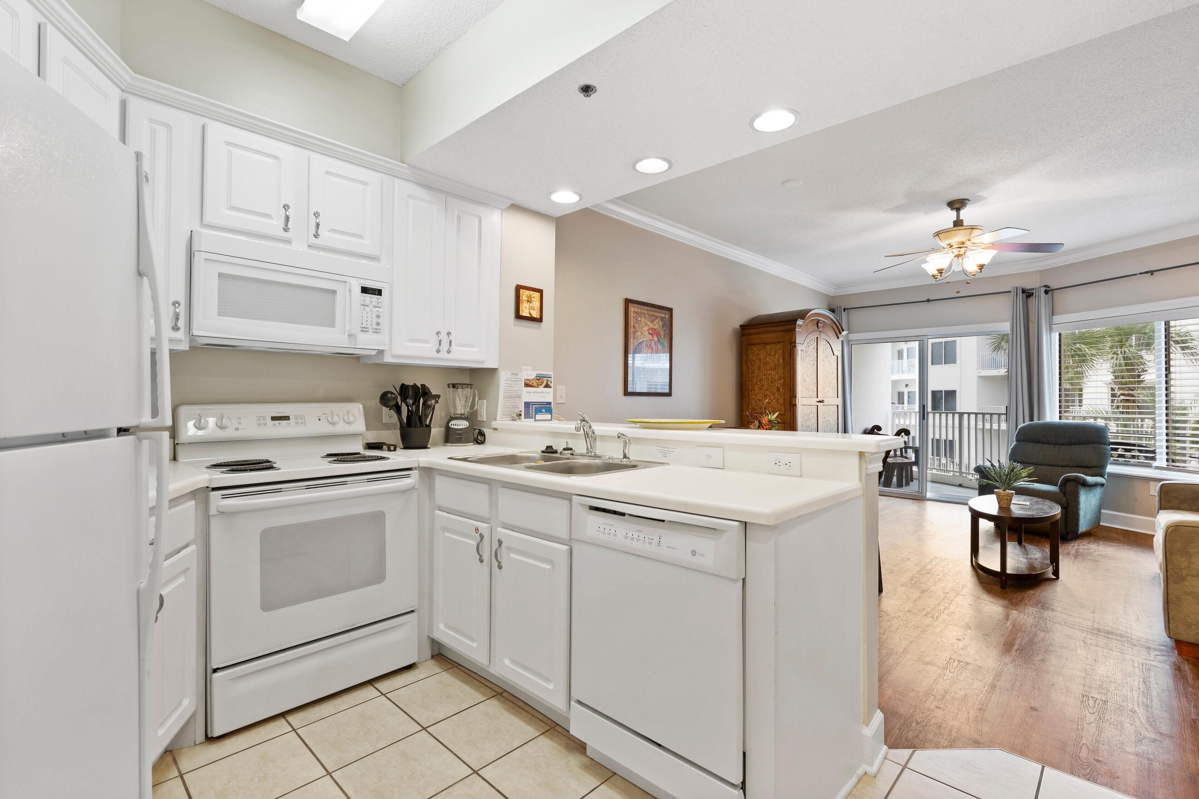 A modern kitchen with white cabinetry, a built-in microwave, and a dishwasher. There is a large island with a sink, and the room has a ceiling fan and large windows.