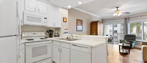 A modern kitchen with white cabinetry, a built-in microwave, and a dishwasher. There is a large island with a sink, and the room has a ceiling fan and large windows.