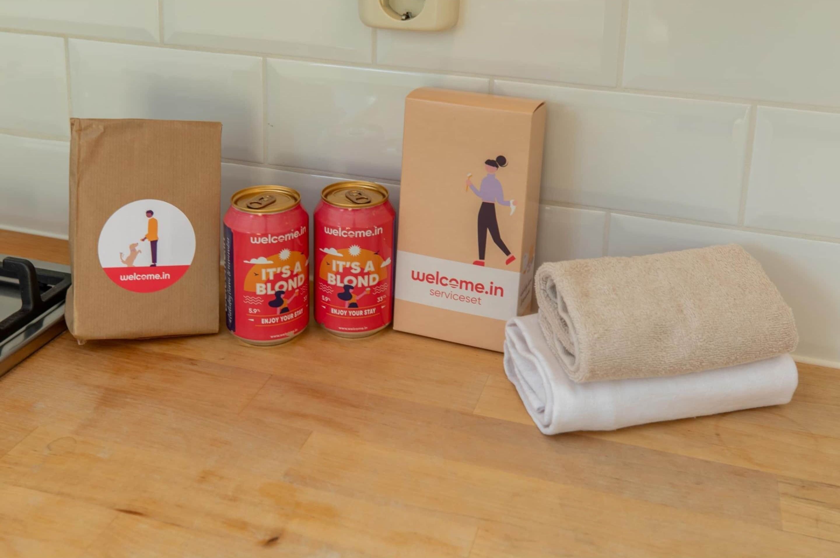 A kitchen counter with a brown paper bag, two cans of 'It's a Blond' drink, a cardboard box with 'Welcome.in' branding, and a folded beige towel.