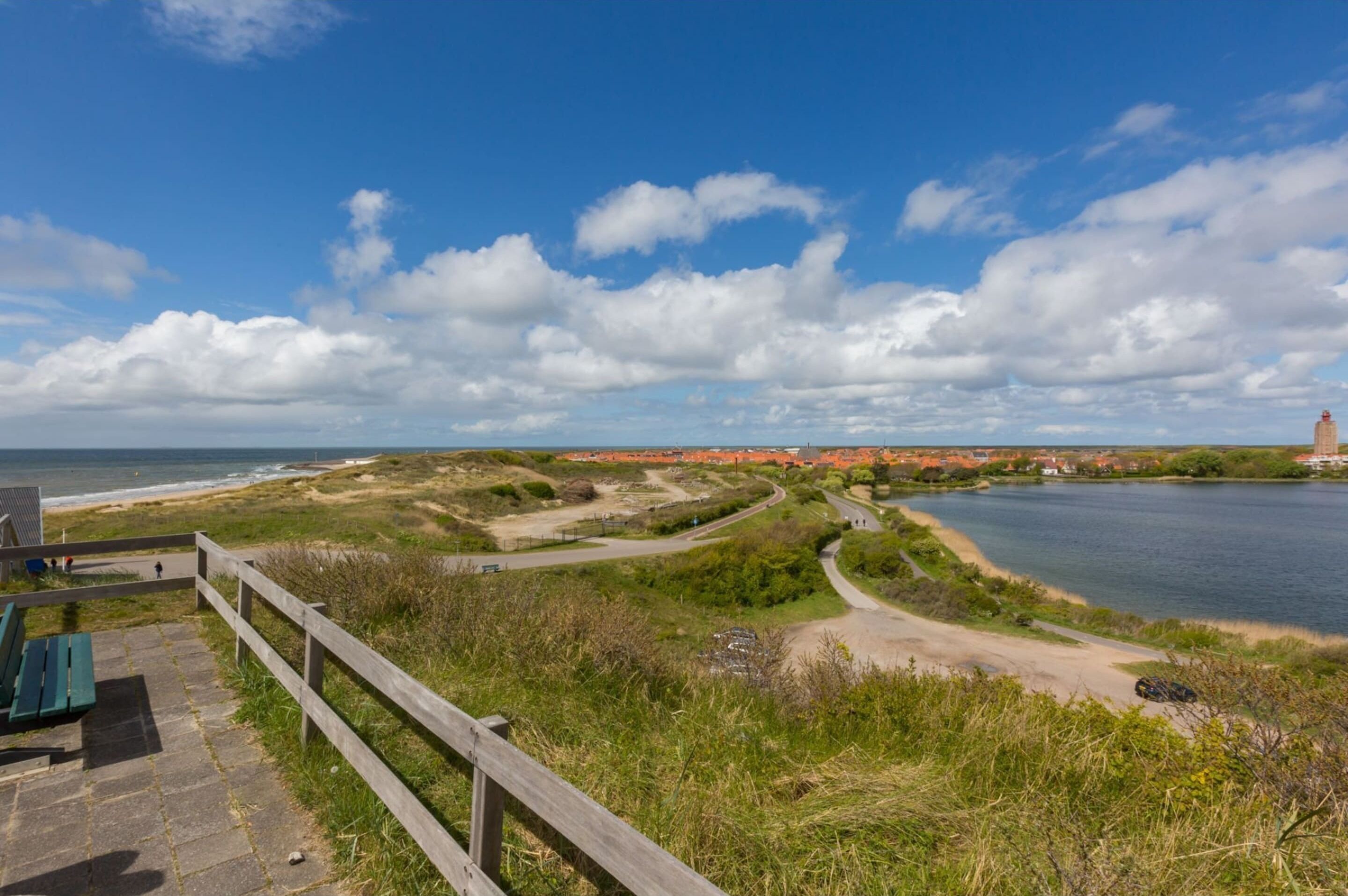 A coastal landscape with a wooden walkway, a bench, and a distant view of a town by the sea.