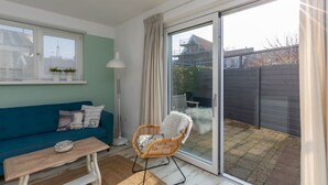 A room with a teal sofa, a wicker chair, a wooden table, and a large sliding glass door leading to a garden. - Holiday Home Near Beach With Sunny Terrace (Westkapelle)