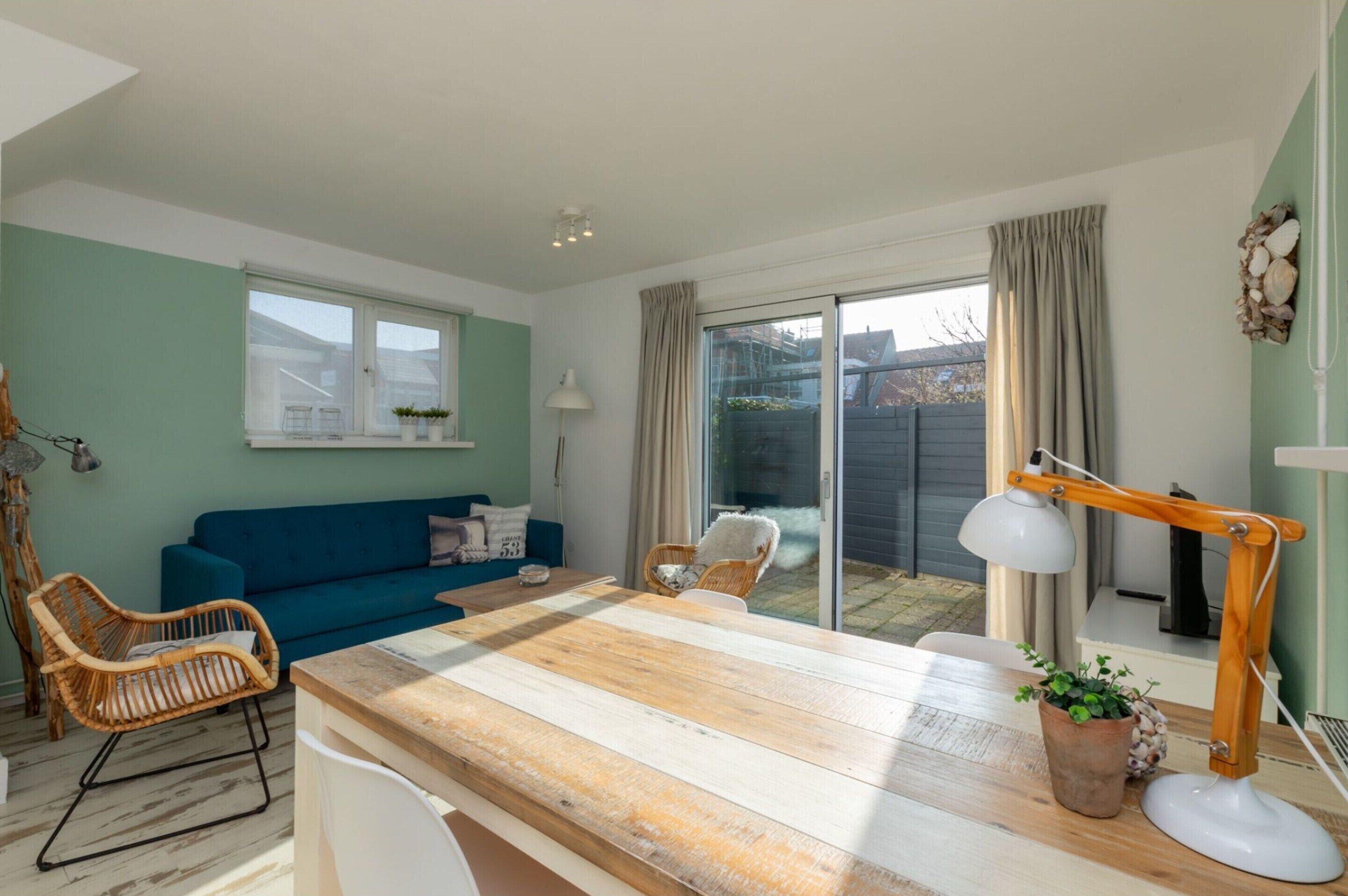 A cozy living room with a blue sofa, wooden coffee table, wicker chairs, and a large window with curtains.