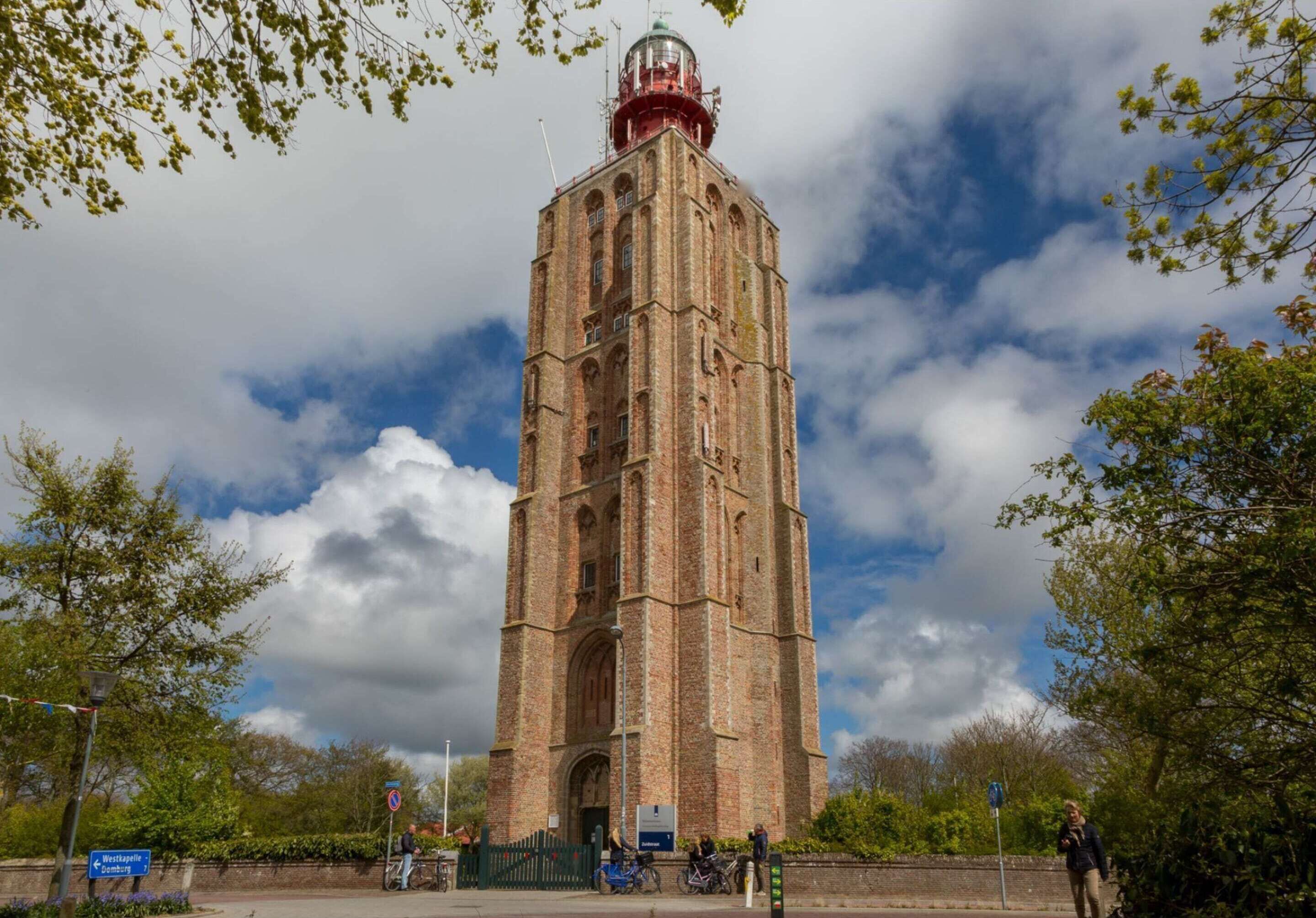 A tall brick tower with a red and white top, surrounded by trees and a partly cloudy sky.