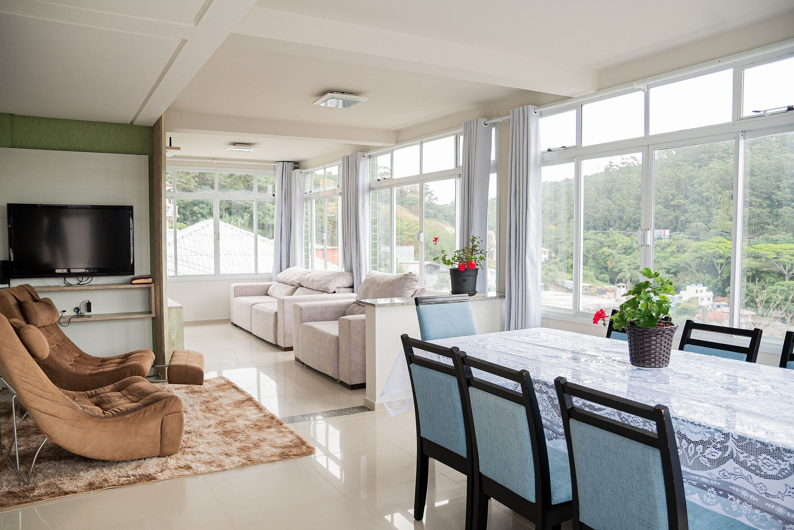 A modern living room with a large window, a flat-screen TV, a sofa, a dining table with chairs, and a massage chair.