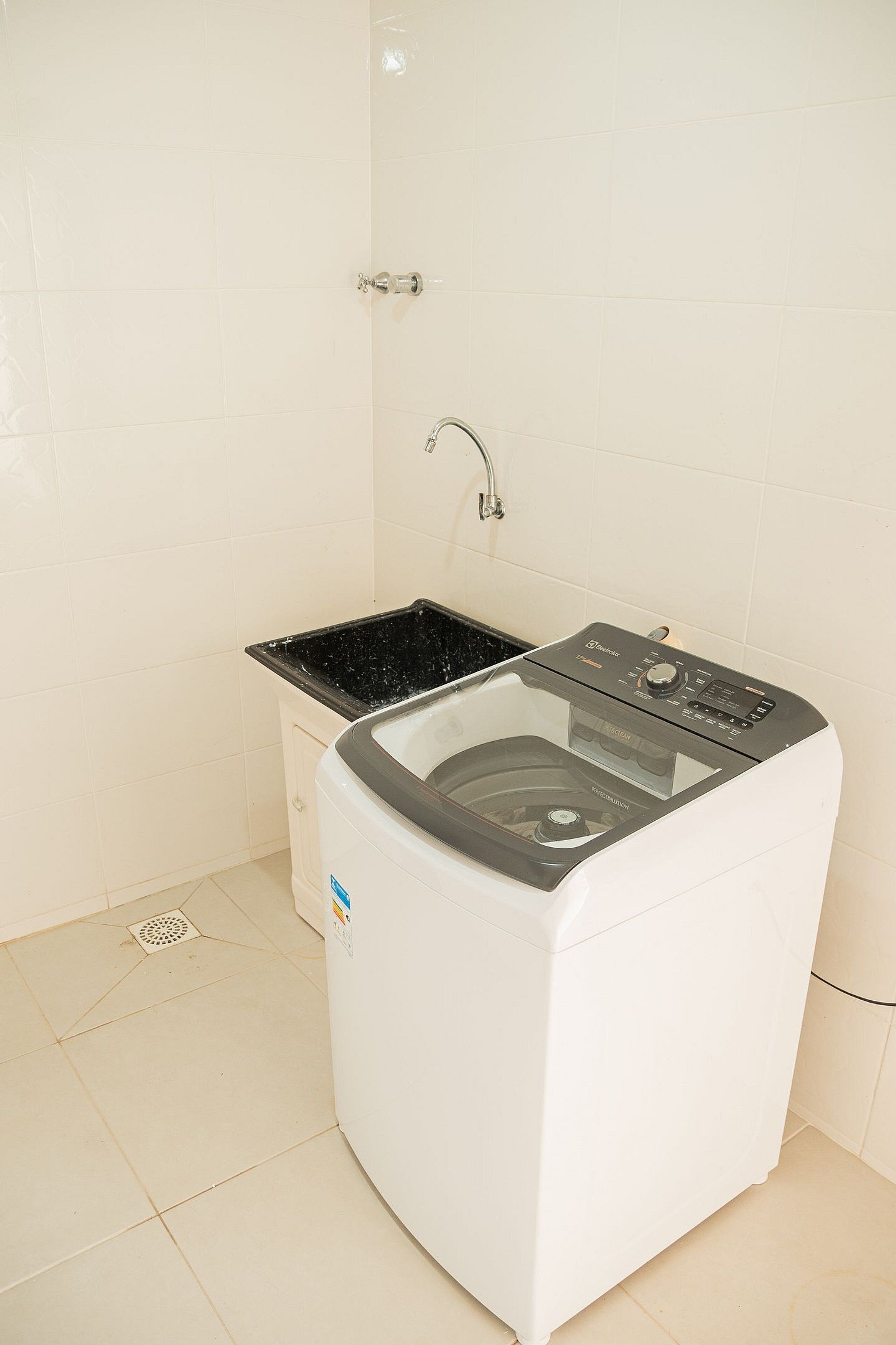 A compact, white washing machine with a black top, placed in a small laundry area with white tiled walls and a stainless steel faucet.