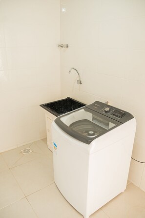 Un lave-linge compact, de couleur blanche, avec un dessus noir, installé dans un petit espace buanderie aux murs recouverts de carreaux blancs et doté d’un robinet en acier inoxydable.