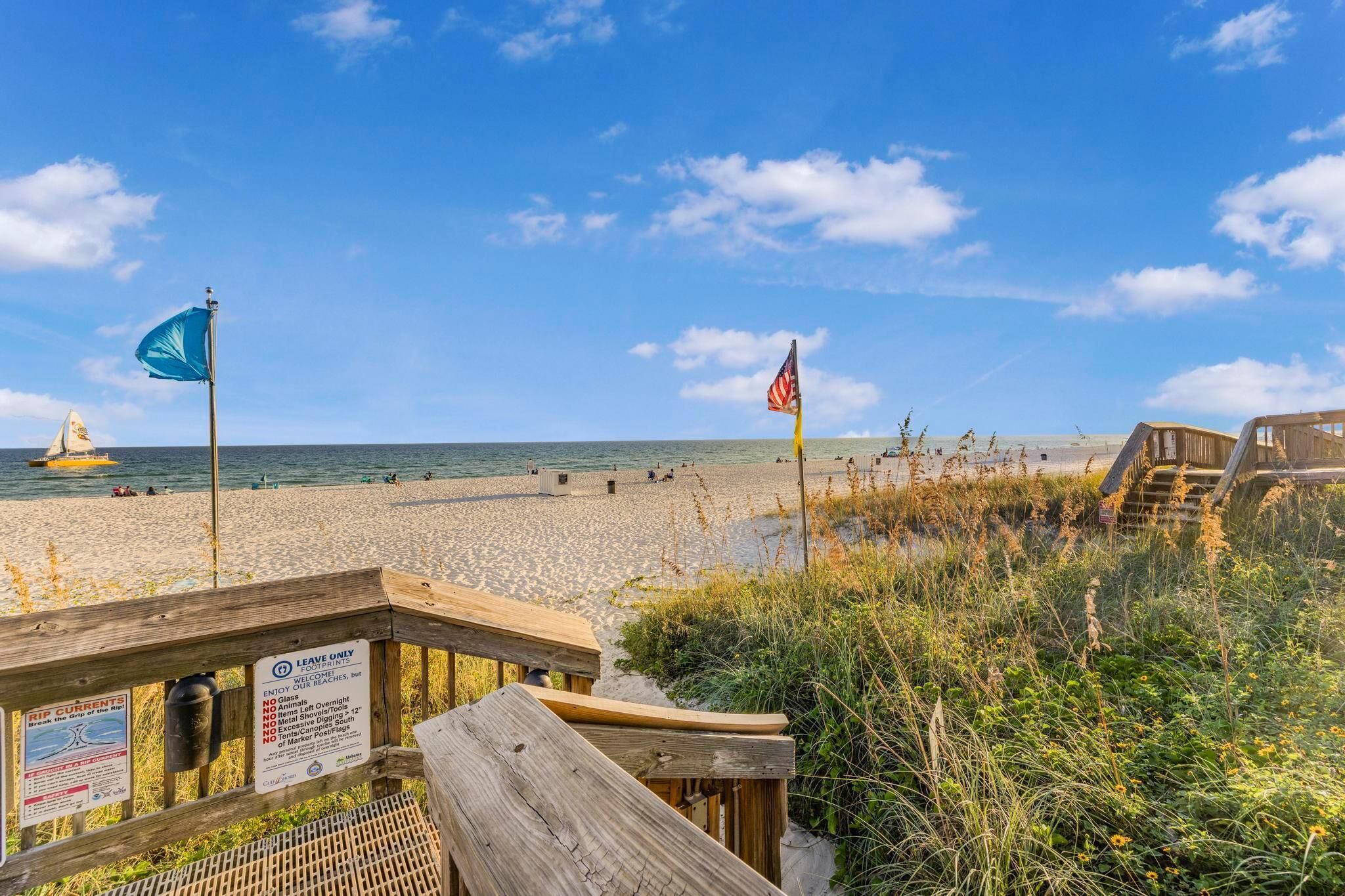 A beach with a wooden boardwalk, a sign indicating beach rules, and a sailboat in the distance.