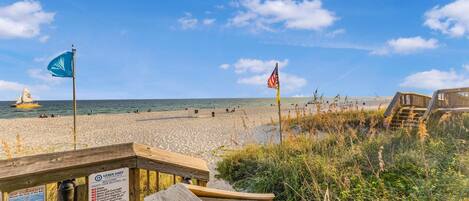 A beach with a wooden boardwalk, a sign indicating beach rules, and a sailboat in the distance.