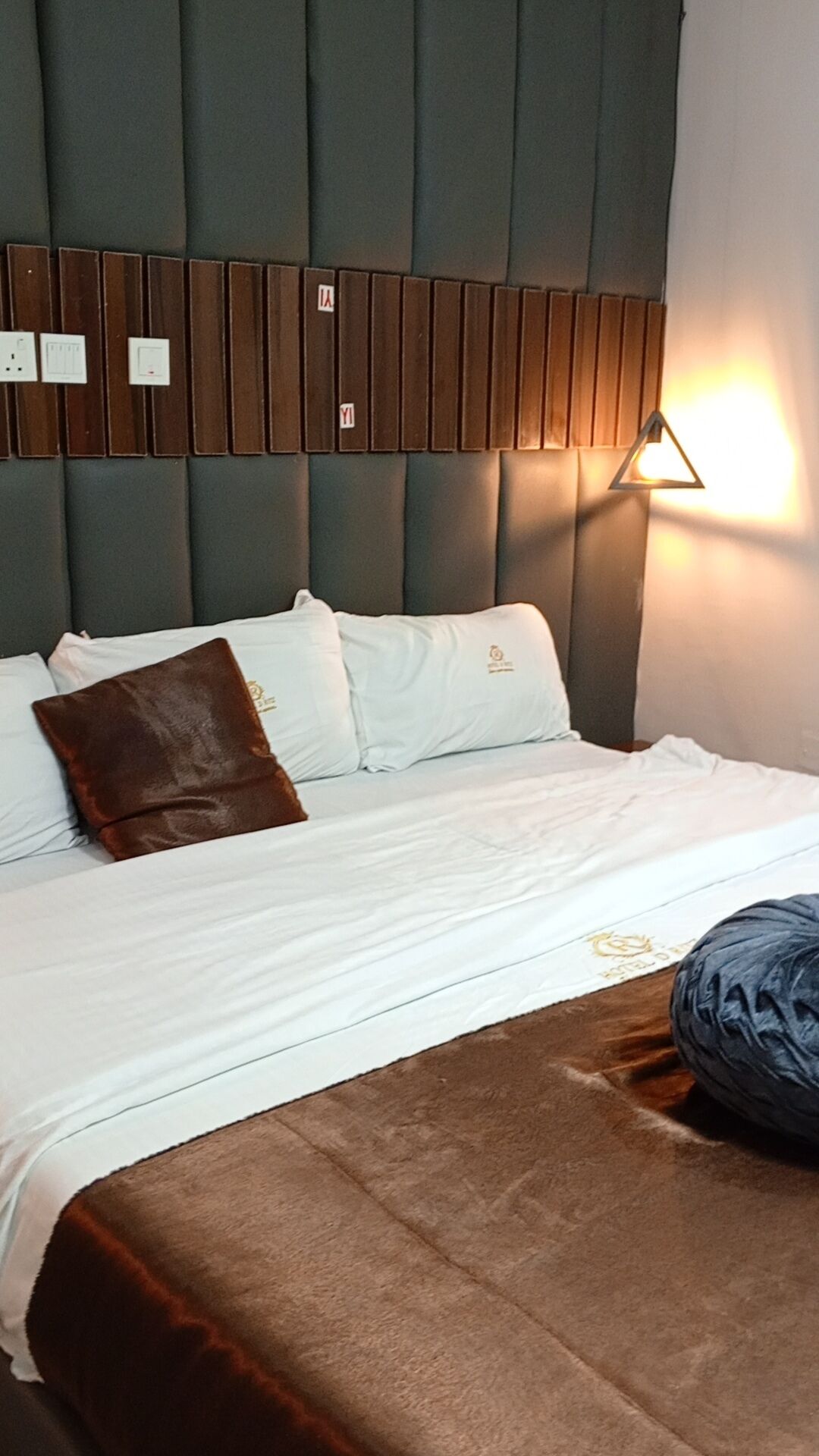 A hotel room with a bed, pillows, a blanket, and a wall-mounted light fixture.