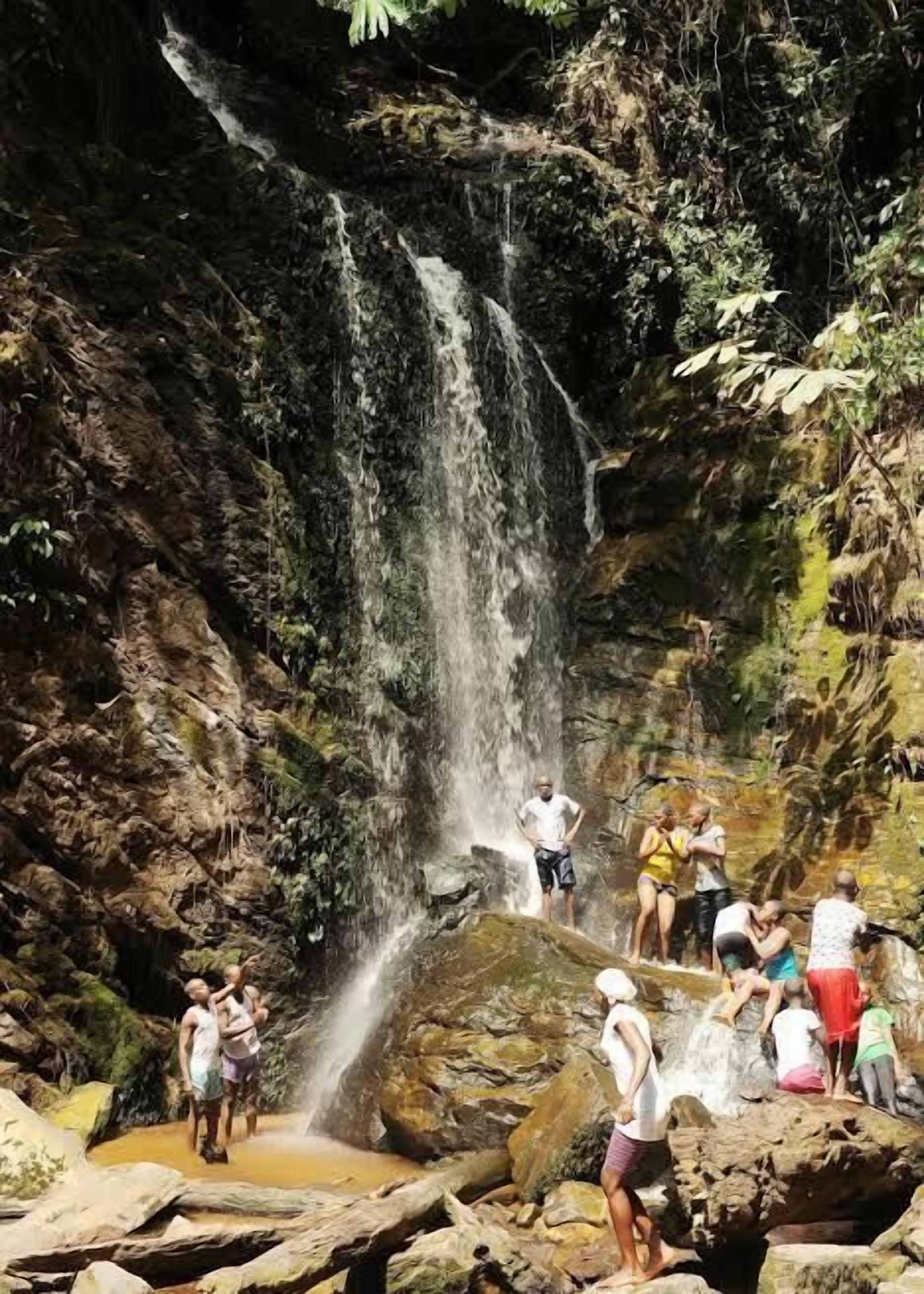A waterfall surrounded by rocks and greenery, with people standing and sitting around it.