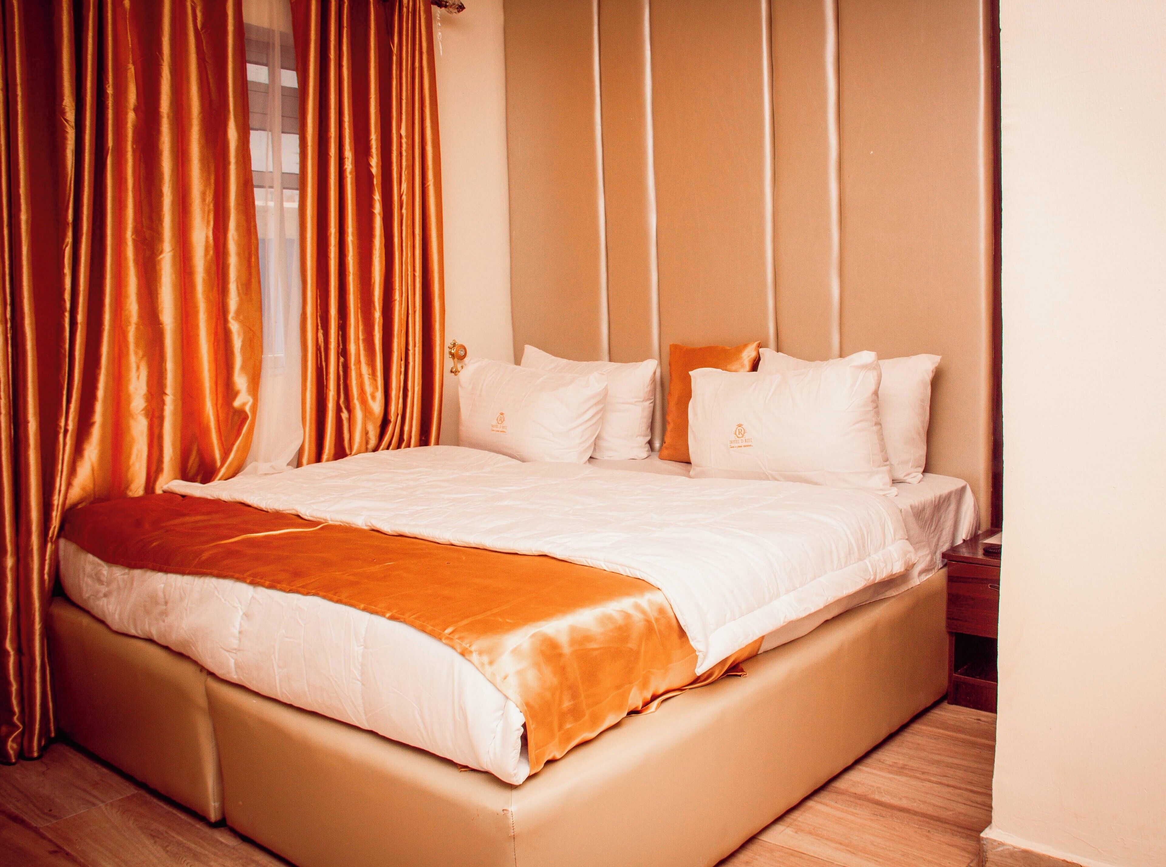 A neatly made bed with white linens and a brown throw, a wooden headboard, and a window with curtains.