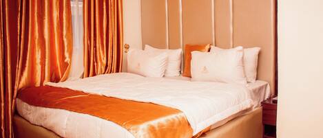A neatly made bed with white linens and a brown throw, a wooden headboard, and a window with curtains.