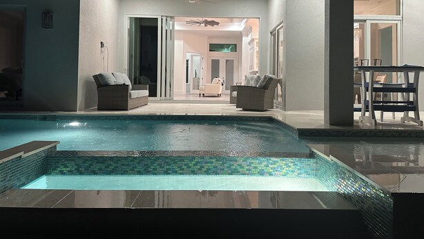 Una piscina con borde de azulejos, un ventilador de techo y un comedor con mesa y sillas.