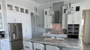 Una cocina moderna con armarios blancos, un refrigerador de acero inoxidable, una isla de mármol con taburetes altos e iluminación colgante.