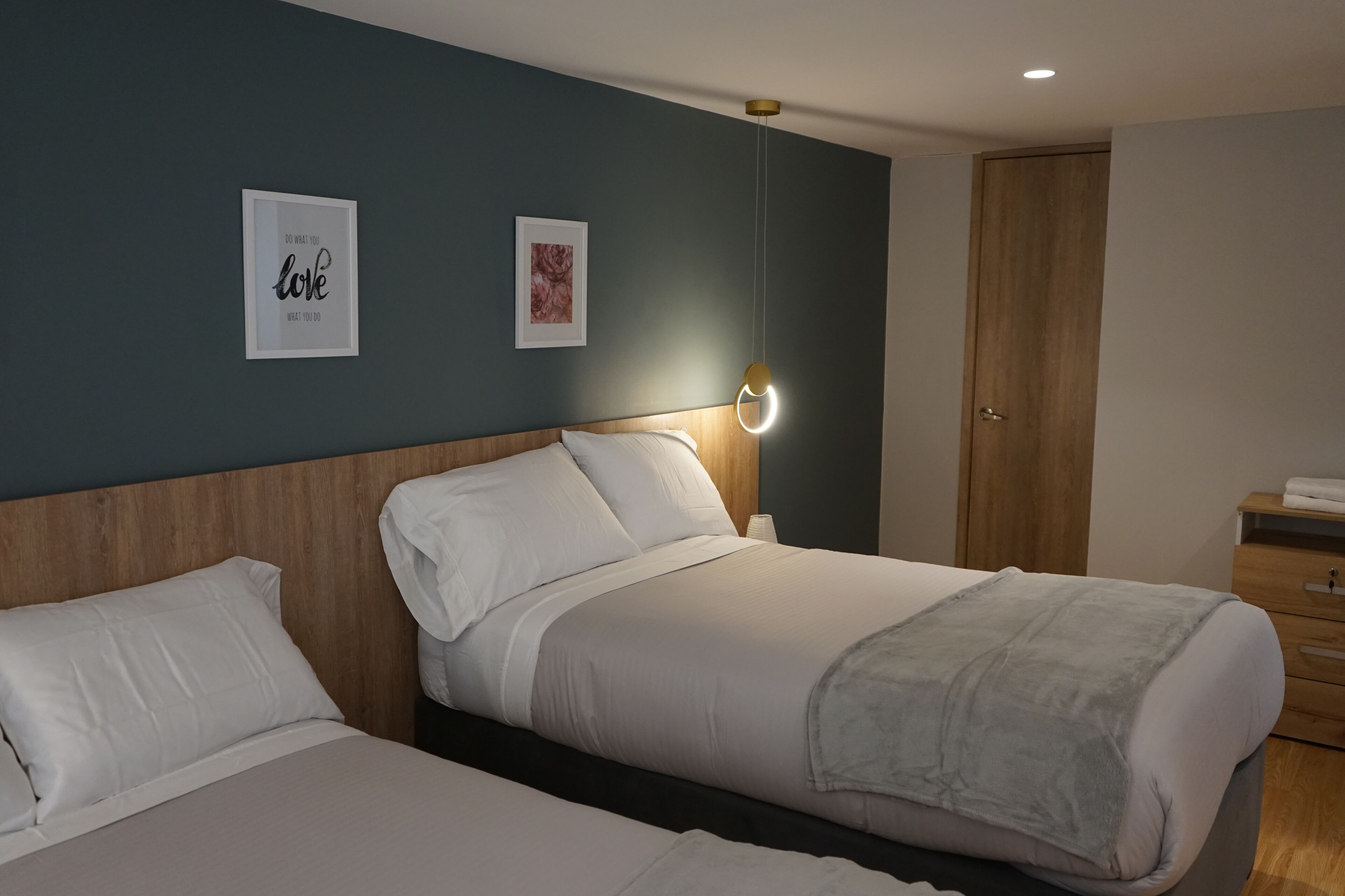Habitación de hotel con dos camas, cabecera de madera, mesita de noche con lámpara y dos cuadros enmarcados en la pared.