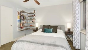 Ein Schlafzimmer mit Bett, Nachttischen, Deckenventilator, Bücherregal und einem Fenster mit Vorhängen.