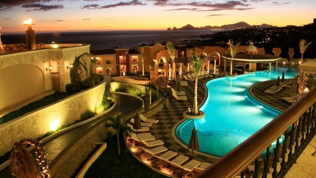 Zona de piscina en un resort con vista al océano al atardecer, rodeada de edificios del resort y palmeras.