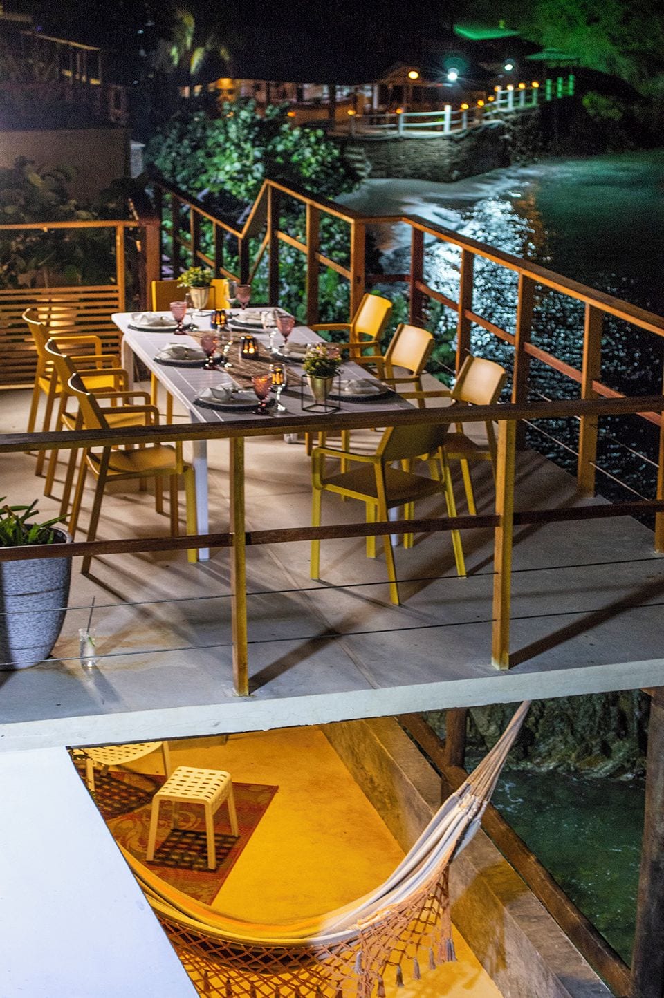 Un espace repas avec une longue table dressée pour quatre personnes, entourée de chaises jaunes, sur une terrasse en bois dotée d’une rambarde.
