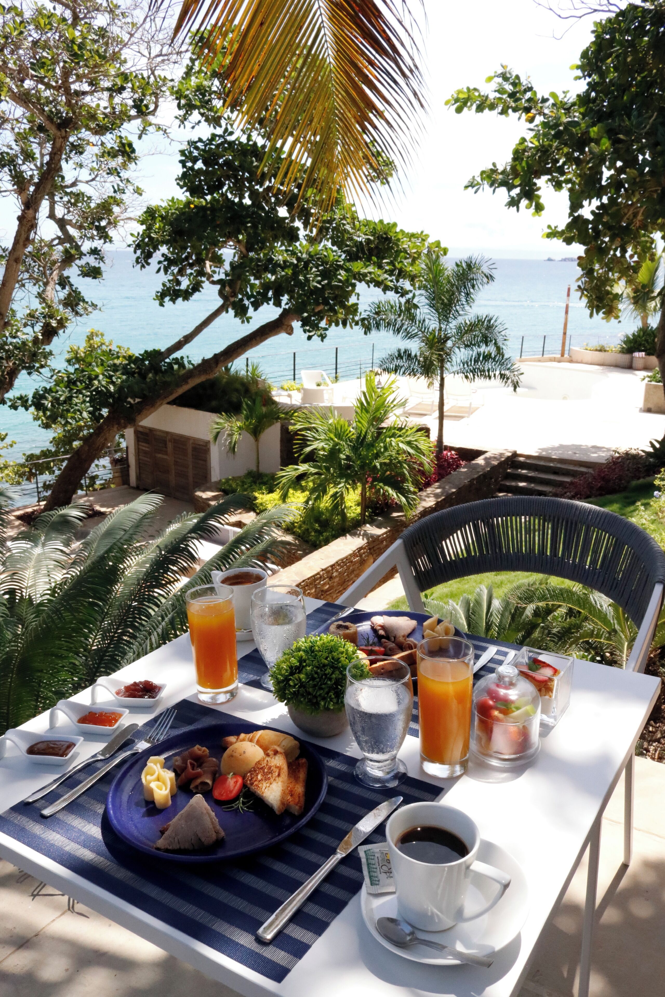 Una tavola imbandita con prodotti per la colazione, tra cui pane tostato, frutta e bevande, con vista su un panorama mozzafiato di mare e palme.