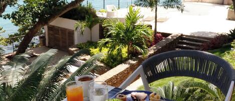 Una mesa dispuesta para el desayuno, con tostadas, fruta y bebidas, ofreciendo vistas panorámicas del mar y palmeras.