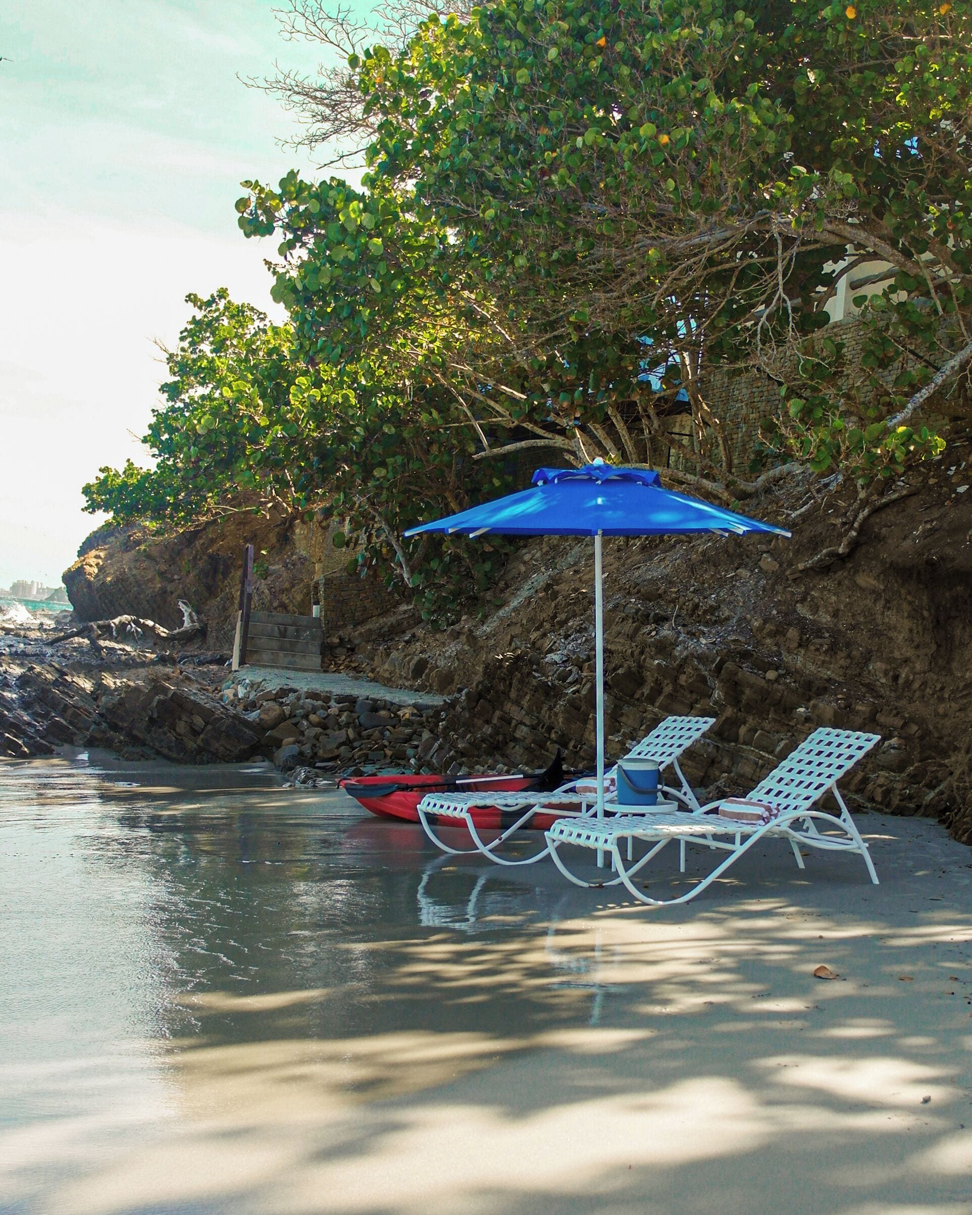 A beachside setting with a blue umbrella, two white lounge chairs, and a red kayak.