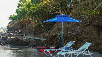 A beachside setting with a blue umbrella, two white lounge chairs, and a red kayak.