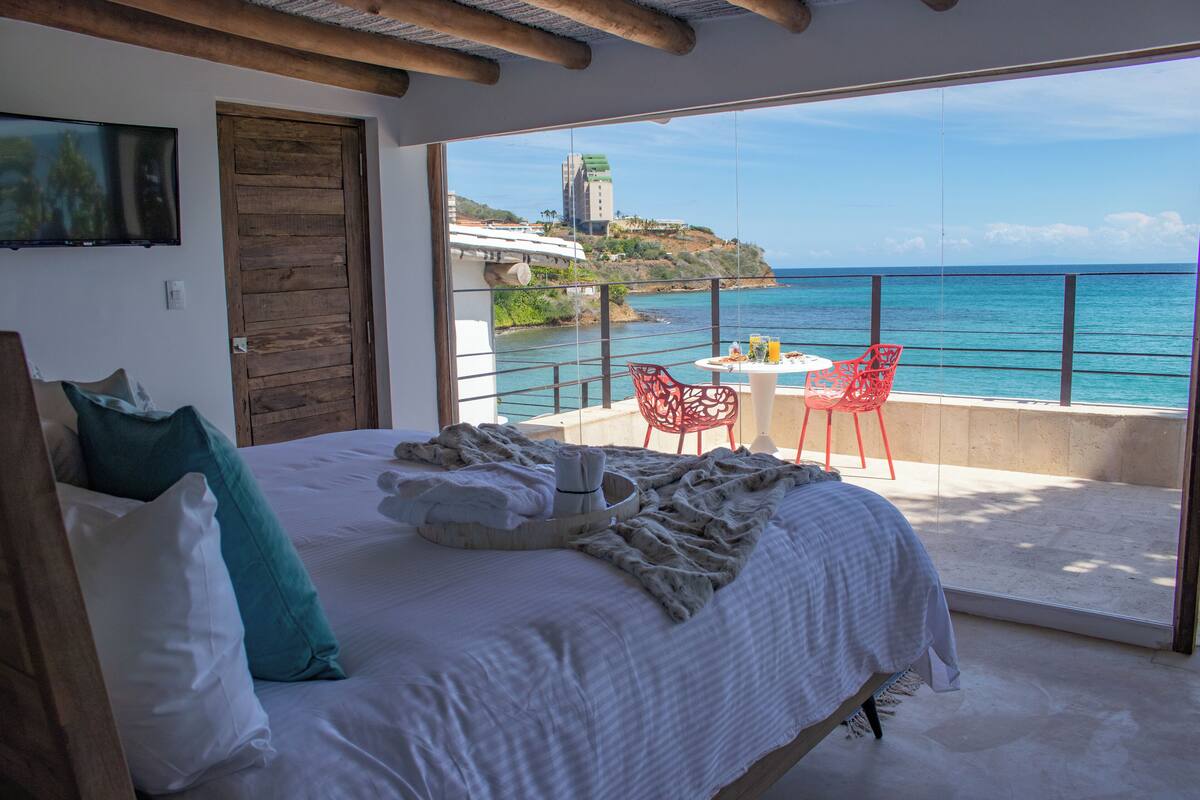 A bedroom with a large bed, a wooden door, and a balcony with a table, chairs, and a view of the ocean.
