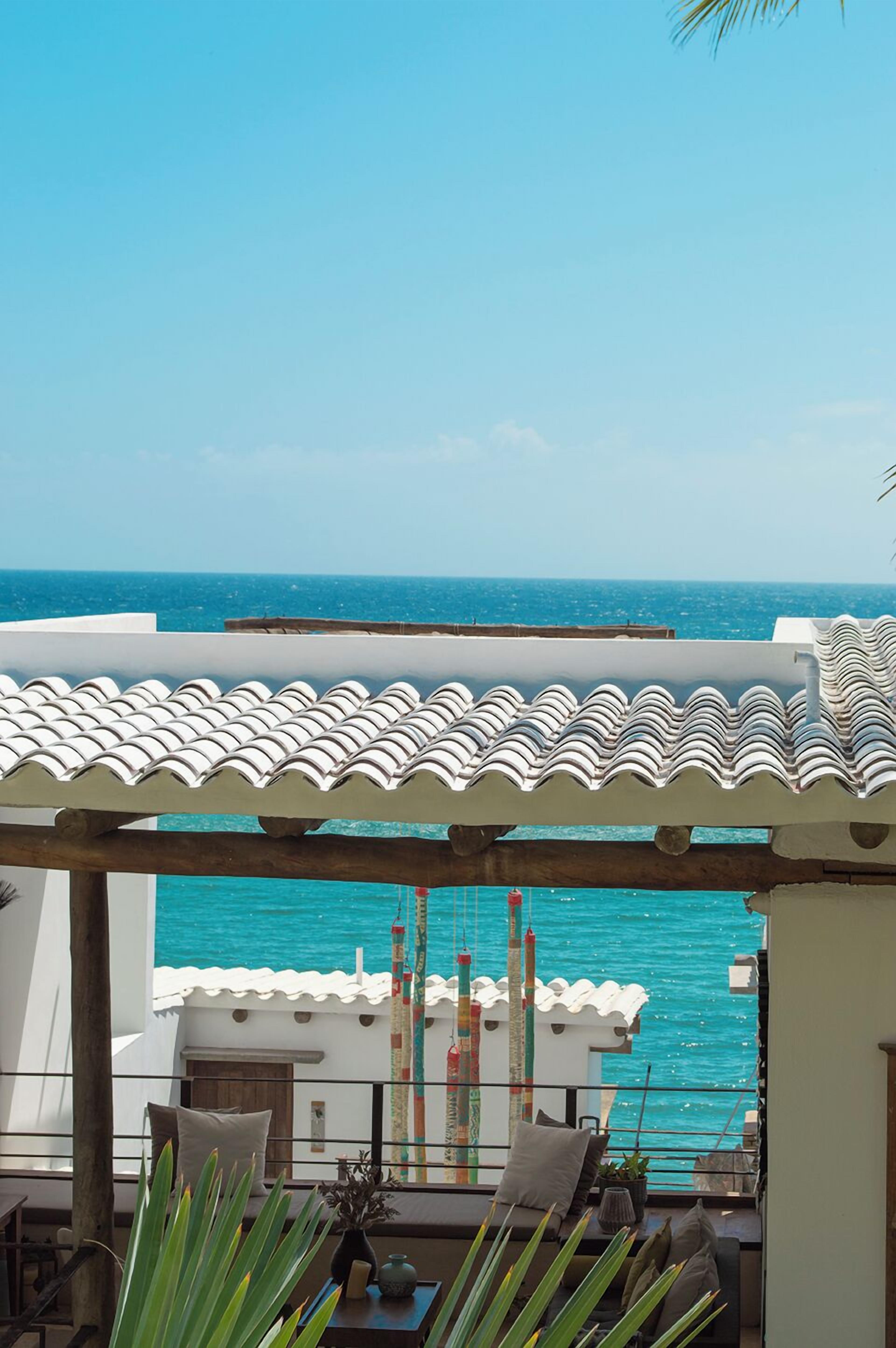 A balcony with a view of the sea, a tiled roof, and a small seating area with cushions and a plant.