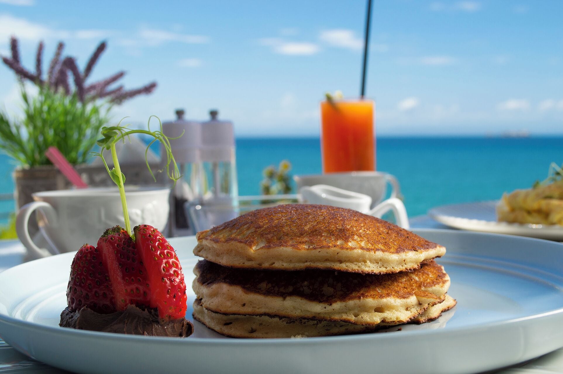 A breakfast setting with pancakes, strawberries, and a glass of orange juice on a table with a view of the ocean.