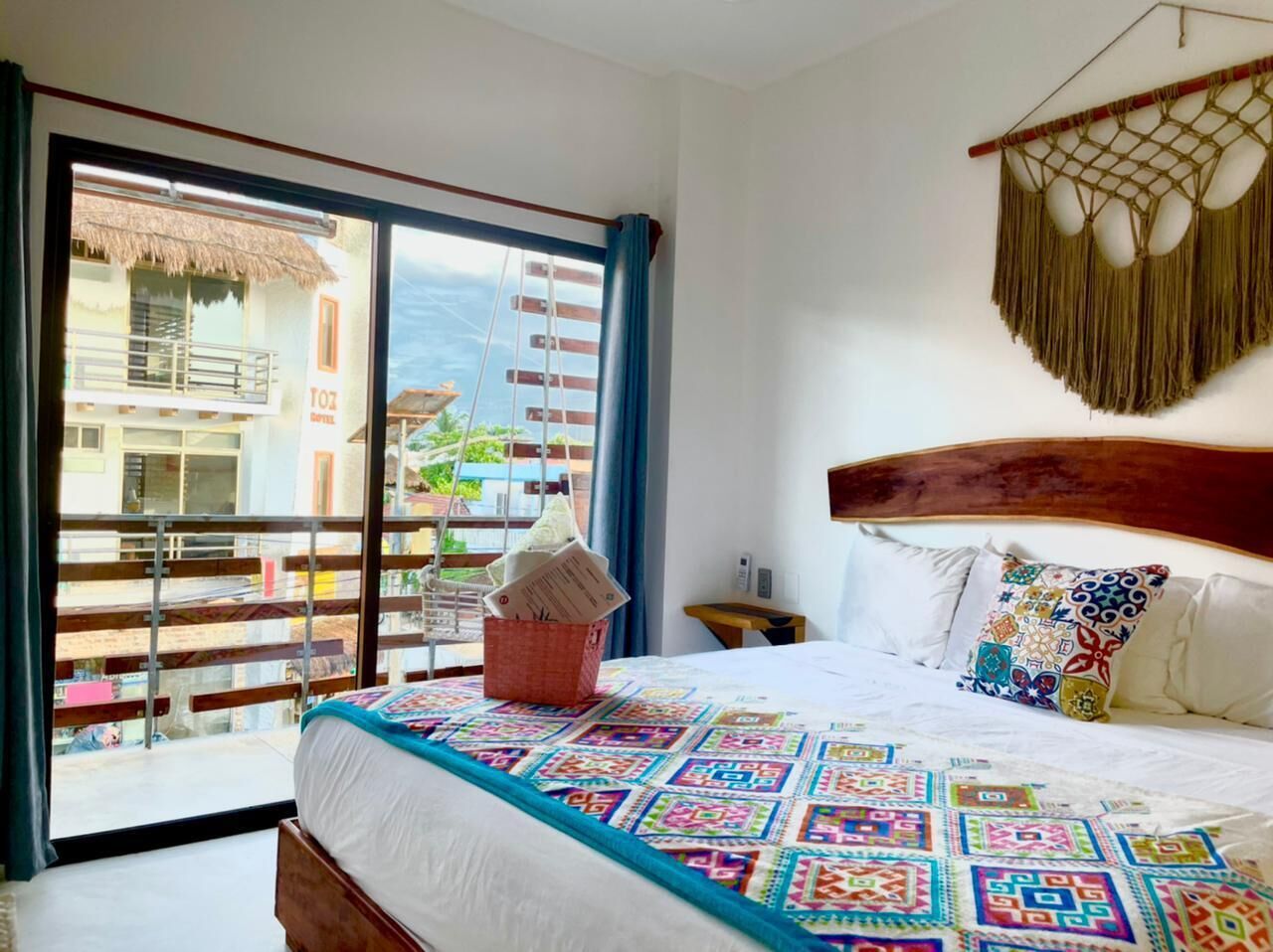 Een hotelkamer met balkon, een bed met kleurrijke quilt, een houten hoofdbord en een hangende macramé decoratie.
