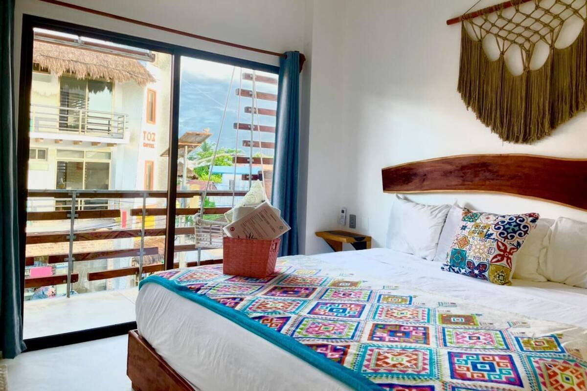 Une chambre d’hôtel avec un balcon, un lit recouvert d’une courtepointe aux couleurs vives, une tête de lit en bois et une décoration murale en macramé.