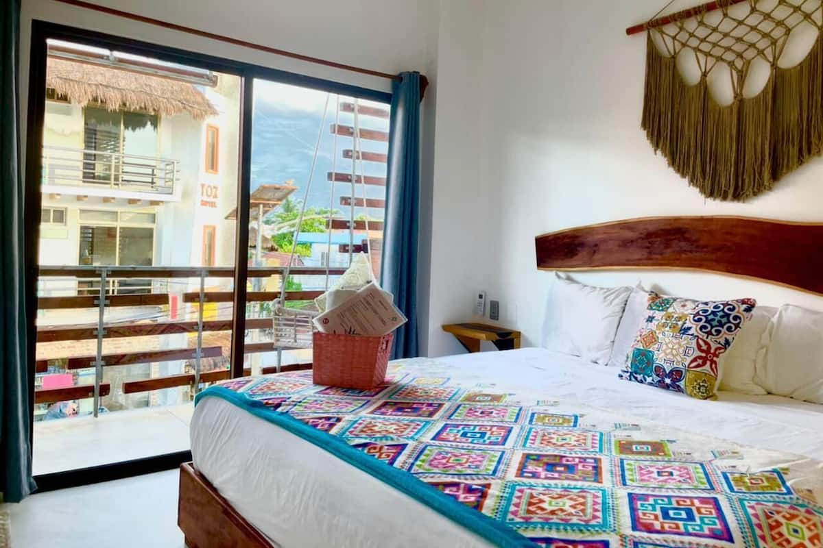 A hotel room with a balcony, a bed with a colorful quilt, a wooden headboard, and a hanging macrame piece.