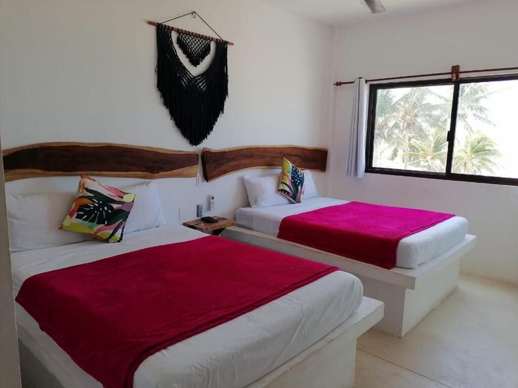 Een kamer met twee bedden, elk met een houten hoofdbord, wit beddengoed en een rode deken. Boven de bedden hangt een decoratie en er is een raam met uitzicht op palmbomen.