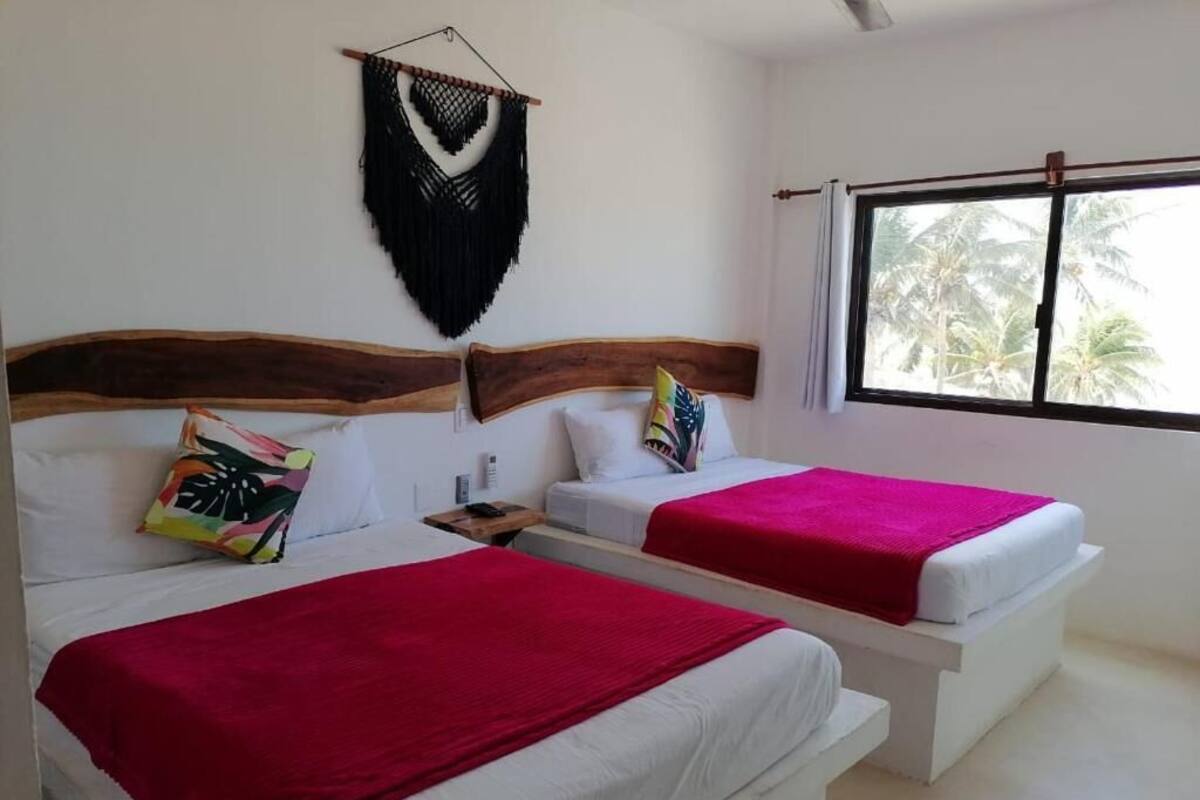 Une chambre avec deux lits, chacun doté d’une tête de lit en bois, de draps blancs et d’une couverture rouge. Un tableau est accroché au mur au-dessus des lits, et une fenêtre offre une vue sur les palmiers.