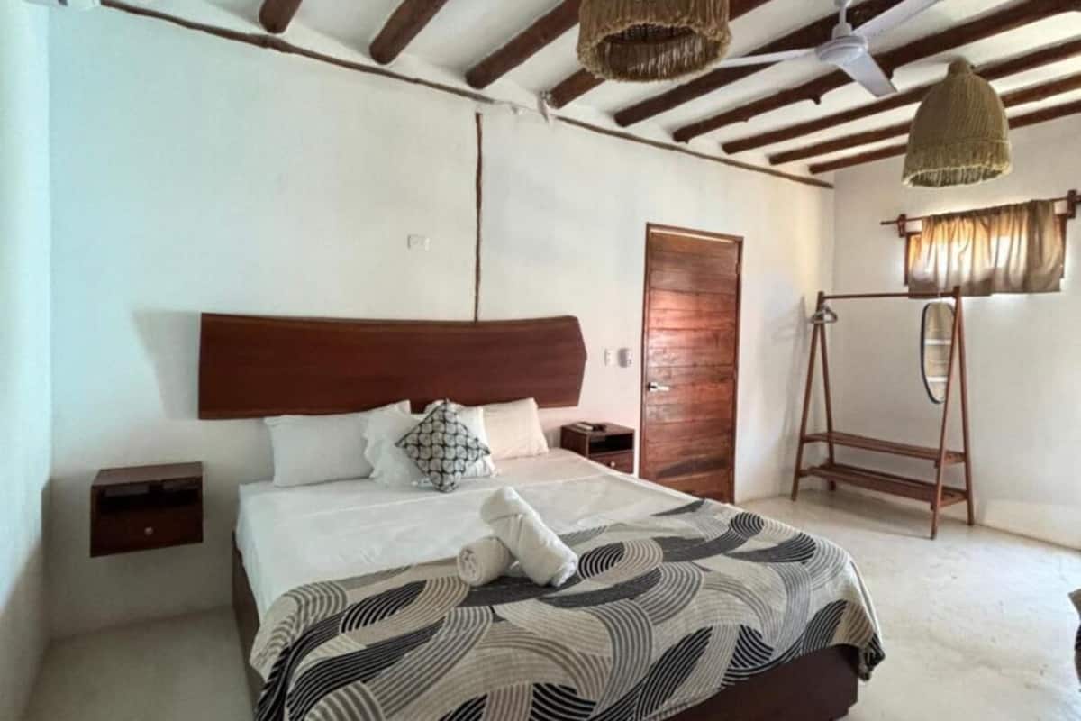 Une chambre d’hôtel avec un grand lit, une tête de lit en bois, une armoire en bois, un radiateur et un ventilateur de plafond.