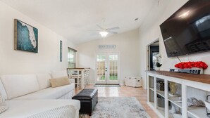 A spacious living area with a white sofa, a coffee table, a TV, and a bookshelf. - Entire JAX Beach Bungalow 7 blocks from Ocean (Jacksonville Beach)