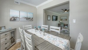 A dining area with a wooden table, white chairs, and a floral table runner. There are three framed pictures on the wall above the table.
