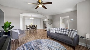 Ein geräumiger Wohnbereich mit grauem Sofa, einem Couchtisch und Deckenventilator. Es gibt eine Essecke mit Tisch und Stühlen, und im Hintergrund ist eine Küche erkennbar.