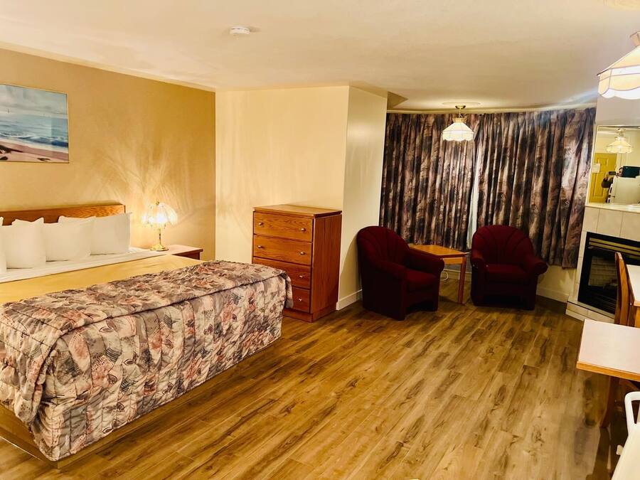 A hotel room with a bed, a wooden dresser, two red armchairs, a small table, and a fireplace.