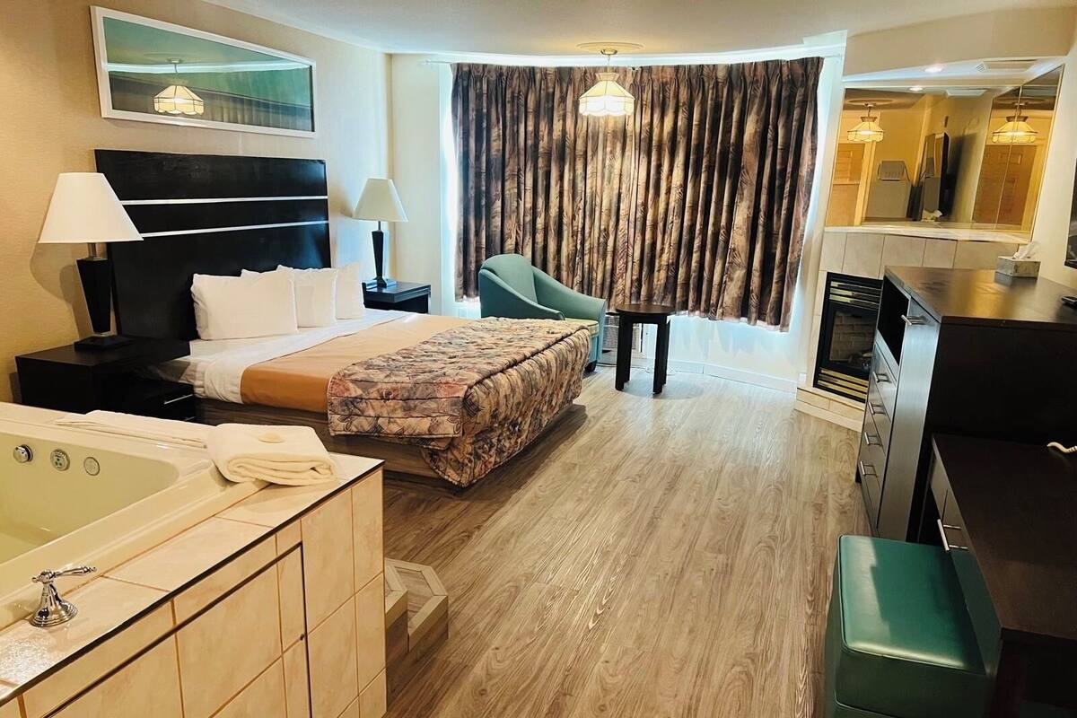 Ein Hotelzimmer mit Bett, Sitzecke mit Sessel, kleinem Tisch, Fernseher, Badezimmer mit Badewanne und Spiegel.