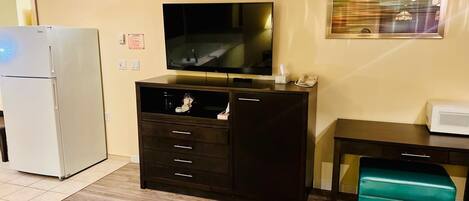 A hotel room with a bed, a television, a minibar, a desk, and a teal ottoman.