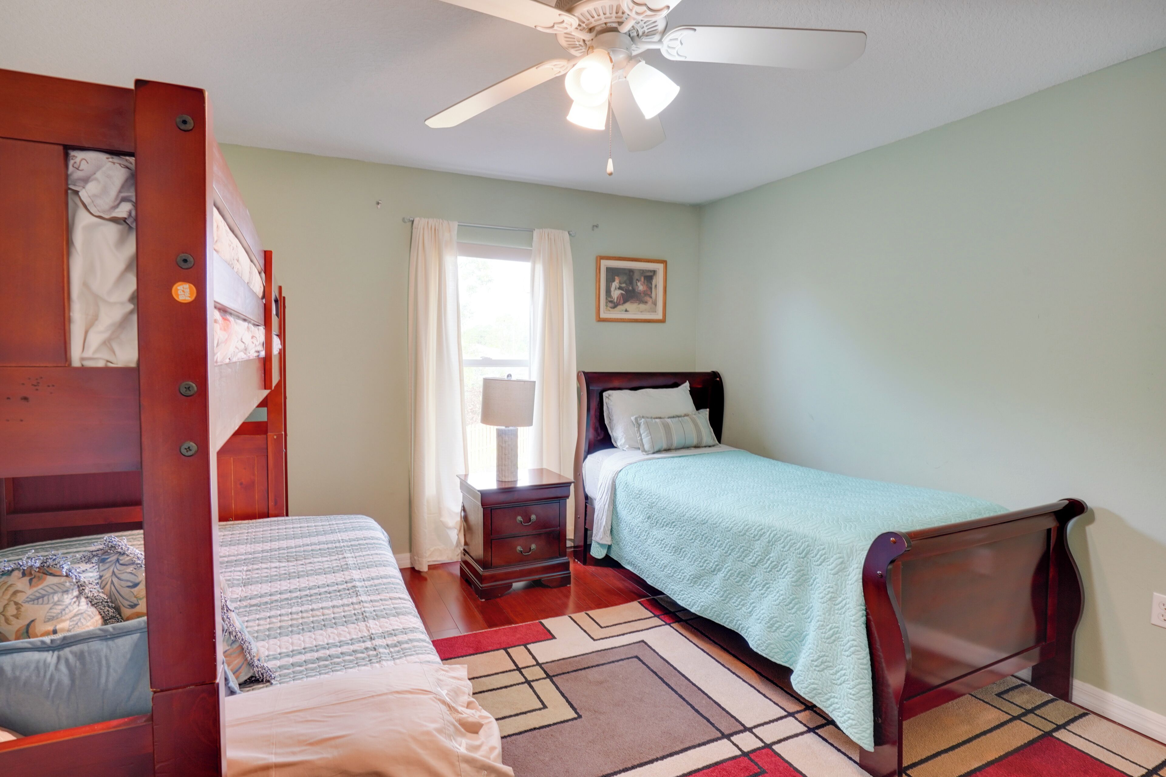 Une chambre avec un lit simple, une table de chevet, une commode, un ventilateur de plafond et une fenêtre avec des rideaux.