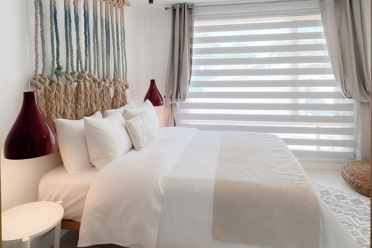 A modern bedroom with a large bed, a bedside table, a red lamp, and a macrame wall hanging.