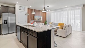 Una cocina moderna con una isla central, electrodomésticos de acero inoxidable, iluminación colgante y un comedor con sofá y cortinas.