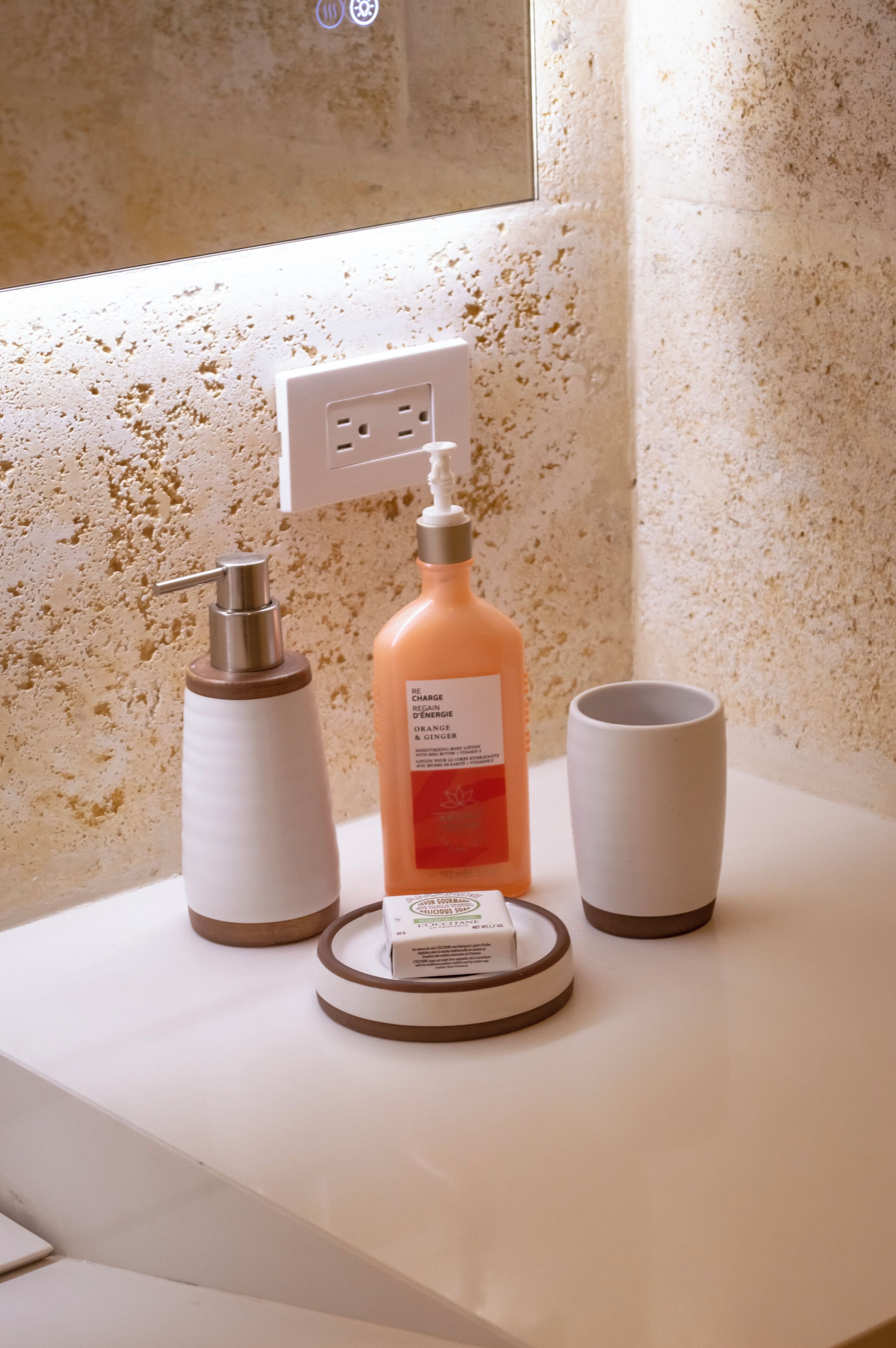 A bathroom counter with a soap dispenser, a bottle of orange body wash, and a cup.