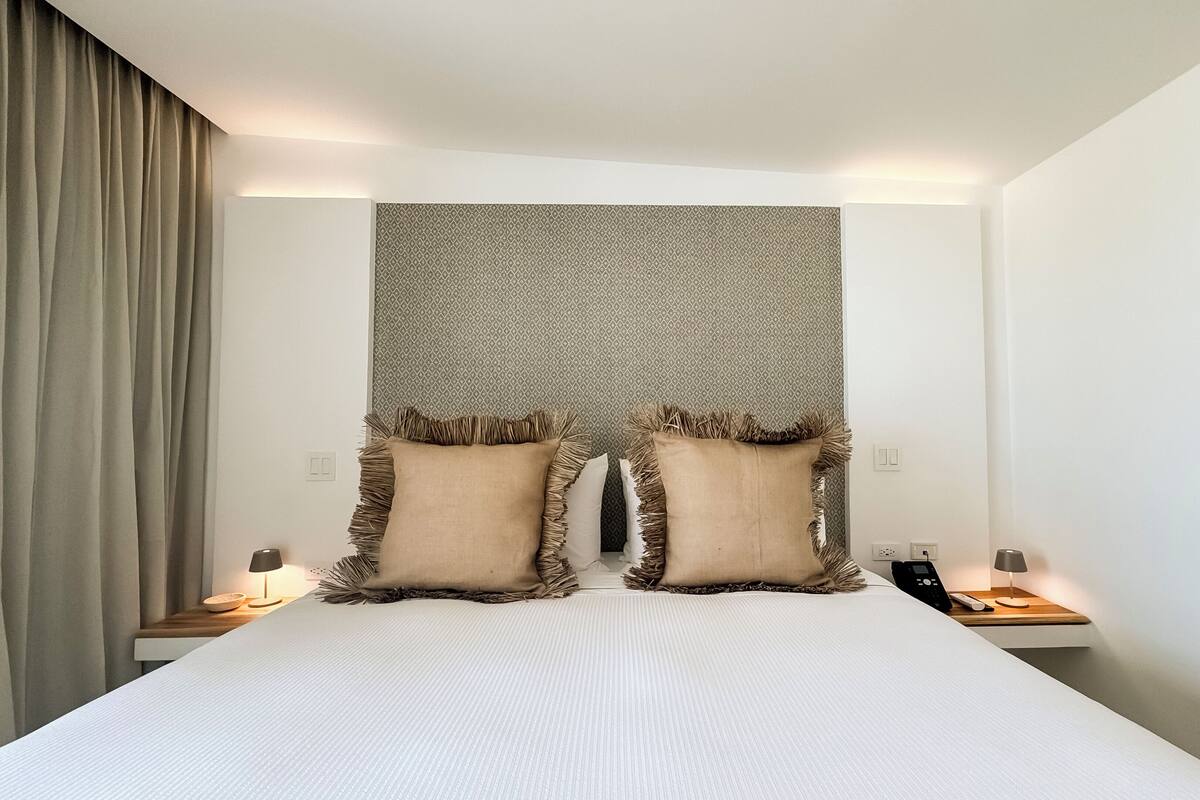 Ein Hotelzimmer mit einem großen Bett, zwei Nachttischen mit Lampen, einer Wand mit Muster und einem Vorhang links.