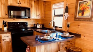 Eine Holzhüttenküche mit einem schwarzen Herd, einer Mikrowelle, einem Toaster, einer Spüle und einem Fenster mit Jalousien. Auf der Kücheninsel stehen zwei Hocker und es liegt eine blaue Unterlage darauf.
